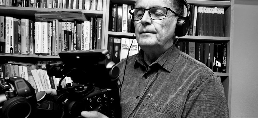 Raoul W. Heimrich mit Kopfhörern an der Kamera vor einem Regal mit Fachbüchern über Drehbuch, Filmrecht und Filmproduktion